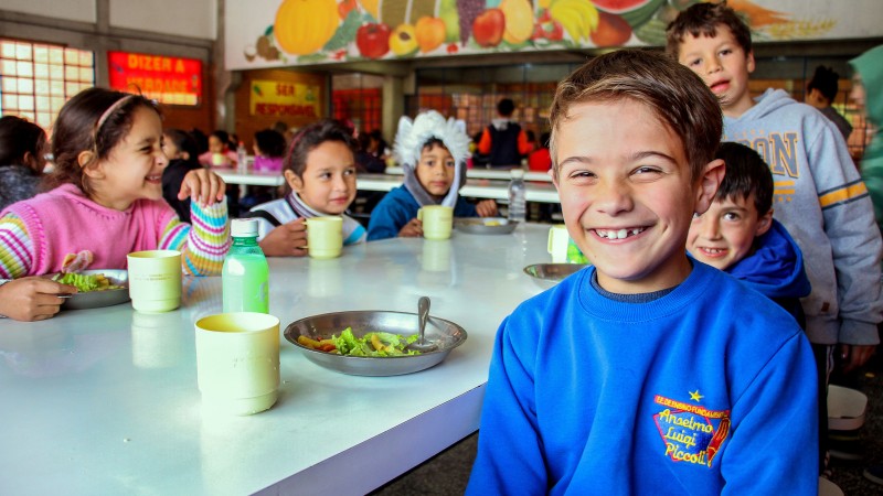 Alunos da Escola Anselmo Luigi Piccoli realizam a merenda no intervalo da aula. Um deles, menino, posa sorridente para foto enquanto os demais se alimentam.