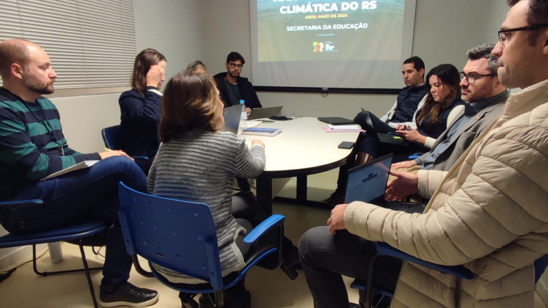 Foto da reunião da Secretaria da Educação com o secretário-executivo adjunto do Ministério da Educação, Gregório Grisa. A equipe está reunida ao redor de uma mesa circular, contando com a presença da secretária estadual de educação, Raquel Teixeira, a secretária-adjunta, Stefanie Eskereski, subsecretários e equipe. Ao fundo da imagem, um slide de uma apresentação diz que se trata da maior catástrofe climática do Rio Grande do Sul. 
