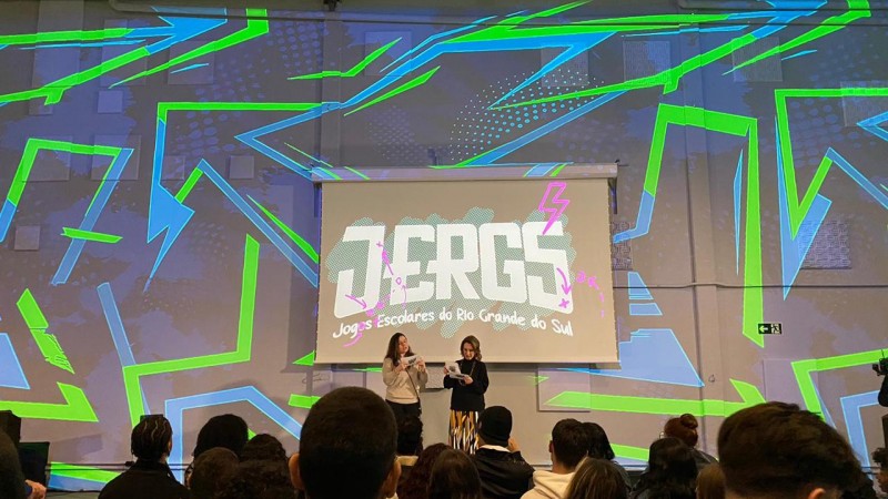 A imagem mostra um auditório moderno, com cores ao fundo em tons de verde e azul. A secretária da educação, Raquel Teixeira, está no centro da foto, apresentando para um grupo de estudantes as ações da Secretaria da Educação. No telão, está escrito, em letras estilizadas, a sigla JERGS, que significa Jogos Escolares do Rio Grande do Sul.