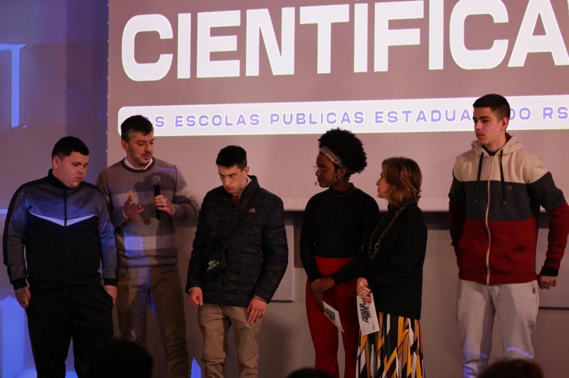 A imagem mostra um grupo de estudantes, enfileirados no palco do evento Nossa Hora. Ao fundo, está um telão com os dizeres Mostra Científica das Escolas Públicas Estaduais do RS.