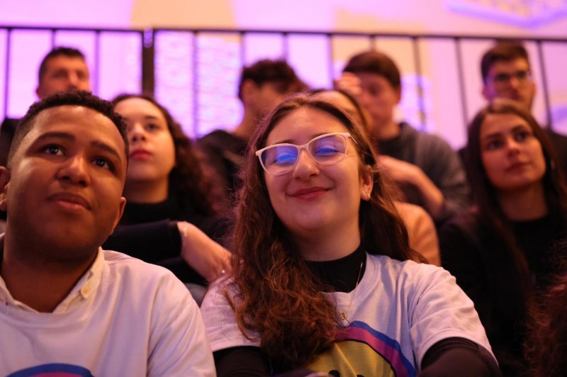 Imagem da plateia do evento Nossa Hora. A foto mostra um rapaz jovem de cabelo curto e pele negra, à esquerda da composição, e uma estudante de pele branca, usando óculos e com cabelos castanhos. Ambos olham atentamente em direção ao auditório, com expressões de sorriso.