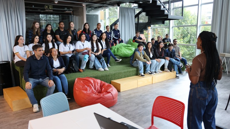 Estudantes e educadores estão sentados em arquibancada de grama sintética, assistindo à fala da diretora da Seduc, Naomy Oliveira, uma mulher de tranças, em um ambiente moderno e iluminado com janelas amplas. Puffs coloridos e um notebook sobre a mesa compõem o espaço.