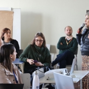 Subsecretária Néri, um mulher loira de altura mediana, está em pé, falando ao microfone diante de um grupo sentado em uma sala de aula. Ela veste casaco preto e saia estampada. Os participantes observam atentamente, com notebooks e projetor sobre a mesa