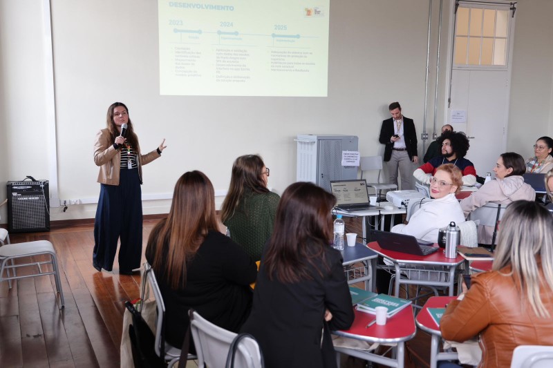 A secretária adjunta da Educação, Stefanie Eskereski, apresenta uma palestra com microfone em mãos para um grupo de profissionais no Instituto de Educação, em Porto Alegre. Participantes acompanham com notebooks e materiais de apoio.