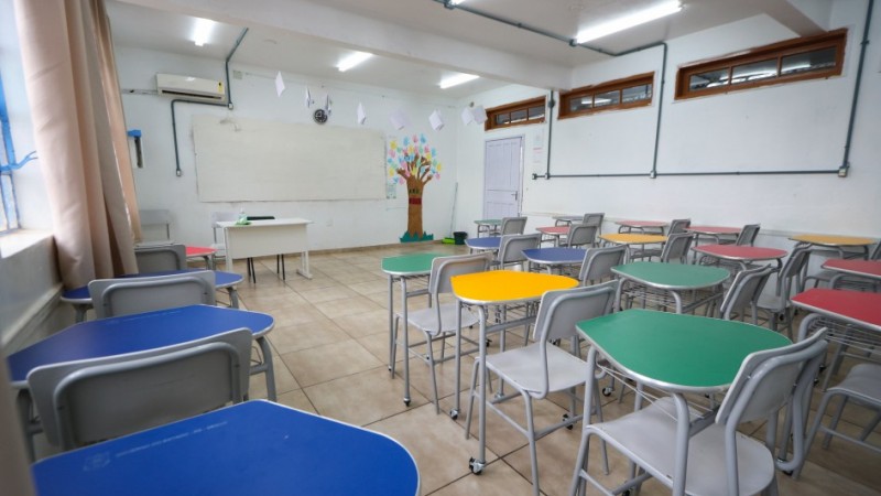 Sala de aula da Escola Dr. Araby Augusto Nácul, com carteiras coloridas organizadas em grupos. Ao fundo, há um quadro branco e um mural com desenho de árvore. Ambiente é bem iluminado, com piso de cerâmica e janelas altas.