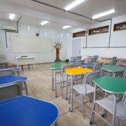 Sala de aula da Escola Dr. Araby Augusto Nácul, com carteiras coloridas organizadas em grupos. Ao fundo, há um quadro branco e um mural com desenho de árvore. Ambiente é bem iluminado, com piso de cerâmica e janelas altas.