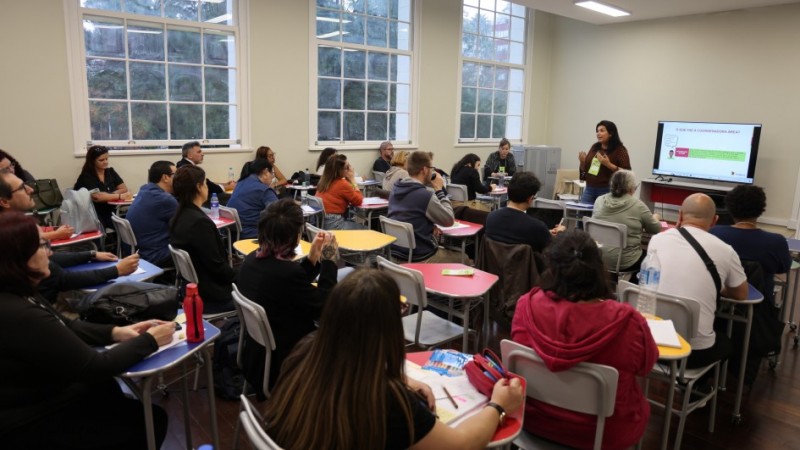 Sala de aula no Instituto de Educação General Flores da Cunha, em Porto Alegre, com pessoas sentadas em mesas coloridas, assistindo a uma apresentação feita por uma mulher em pé, diante de uma tela grande. Ao fundo, há três janelas amplas.