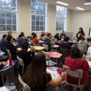 Sala de aula no Instituto de Educação General Flores da Cunha, em Porto Alegre, com pessoas sentadas em mesas coloridas, assistindo a uma apresentação feita por  uma mulher em pé, diante de uma tela grande. Ao fundo, há três janelas amplas.