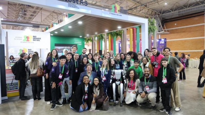 Foto de um grande grupo de pessoas reunidas em frente ao estande do Governo do Rio Grande do Sul, em um evento realizado em um pavilhão coberto. A maioria usa crachás coloridos e colares verdes. Ao centro, há um robô humanoide com braços estendidos, rodeado por pessoas sorridentes, muitas delas jovens. O ambiente tem decoração moderna, com cores vivas, plantas e letreiros.
