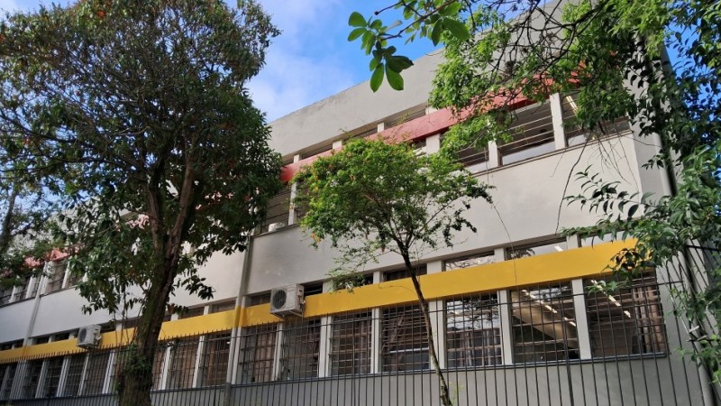 A imagem mostra a fachada da escola, um prédio de dois andares, com janelas grandes e grades, pintado de cinza e amarelo. Há árvores no primeiro plano, e o céu está azul com algumas nuvens