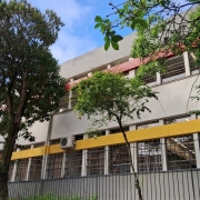 A imagem mostra a fachada da escola, um prédio de dois andares, com janelas grandes e grades, pintado de cinza e amarelo. Há árvores no primeiro plano, e o céu está azul com algumas nuvens