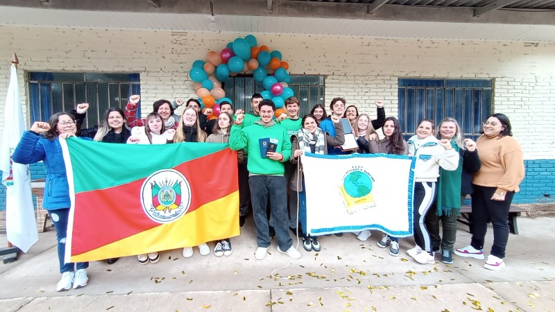  Grupo de estudantes e educadores posa sorridente em frente a uma escola, segurando bandeiras do Rio Grande do Sul e da Conferência Infantojuvenil pelo Meio Ambiente. Ao fundo, há uma parede com janelas e uma decoração de balões coloridos. Todos levantam o punho em sinal de celebração.