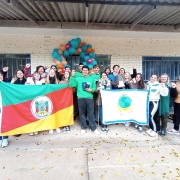  Grupo de estudantes e educadores posa sorridente em frente a uma escola, segurando bandeiras do Rio Grande do Sul e da Conferência Infantojuvenil pelo Meio Ambiente. Ao fundo, há uma parede com janelas e uma decoração de balões coloridos. Todos levantam o punho em sinal de celebração.