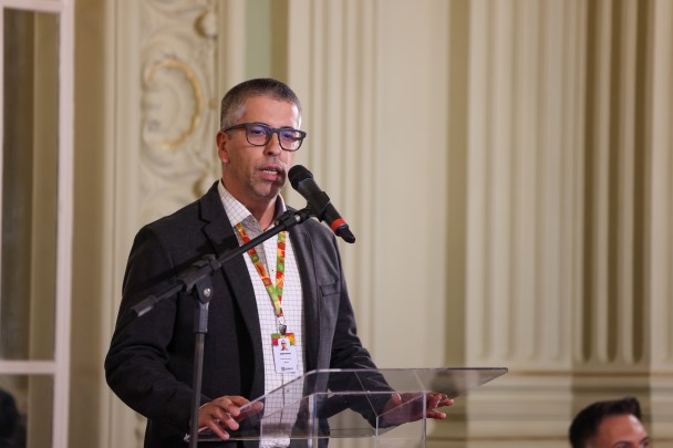 O ChatGPT disse:
Homem de óculos fala ao microfone em púlpito transparente durante evento. Ele usa crachá e paletó escuro, com fundo de colunas e ornamentos clássicos.
