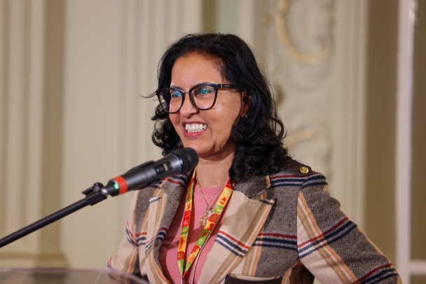 O ChatGPT disse:
Mulher sorri ao falar ao microfone em púlpito transparente. Usa óculos, blusa rosa e casaco xadrez. Ao fundo, parede clara com detalhes ornamentais.
