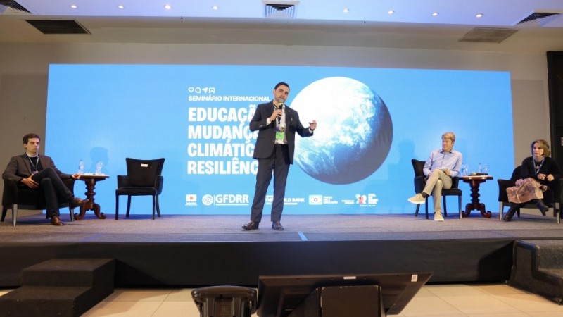 Esta é uma fotografia de um painel de discussão em um seminário. No centro da imagem, em um palco elevado, um homem com barba e vestindo um terno escuro está em pé, no meio de outras três pessoas sentadas. Ele gesticula com as mãos enquanto fala para uma plateia que não está visível. Atrás do palco, um grande telão azul exibe o título "SEMINÁRIO INTERNACIONAL" e as palavras "EDUCAÇÃO, MUDANÇAS CLIMÁTICAS e RESILIÊNCIA", junto com logotipos de organizações como "GFDRR" e "WORLD BANK". No palco, à esquerda do orador, um homem de terno escuro está sentado em uma poltrona preta, com as pernas cruzadas. À direita do orador, um homem de cabelo claro e camisa social está sentado, e ao seu lado, uma mulher de cabelo escuro, vestindo uma blusa preta, também está sentada. O chão do palco é preto e há mesas de apoio com garrafas de água. A iluminação é focada no palco, destacando os participantes.