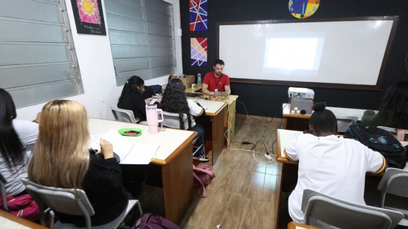 Sala de aula com alunos sentados em carteiras, de frente para um professor que ministra aula com auxílio de projetor. A lousa ao fundo exibe uma projeção. As paredes têm desenhos coloridos e há mochilas sobre as mesas e no chão.