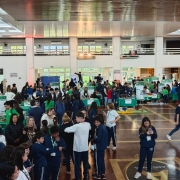 A foto mostra uma feira de ciências em um salão amplo e iluminado, com estudantes interagindo com estandes e pôsteres verdes. O espaço tem piso de madeira brilhante e teto alto.
