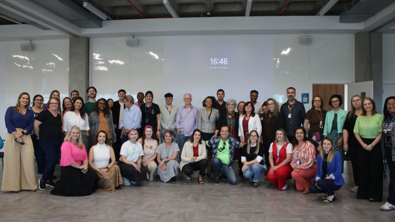 Cerca de 40 pessoas posam para uma foto em grupo em uma sala ampla e iluminada. Algumas est&atilde;o em p&eacute; e outras agachadas na frente. Ao fundo, h&aacute; uma parede branca com um projetor.