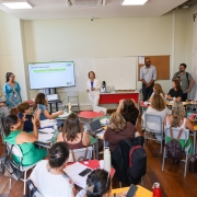 Sala de aula com cerca de 20 educadores sentados em mesas organizadas em c&iacute;rculo. Ao centro, a secret&aacute;ria da Educa&ccedil;&atilde;o, Raquel Teixeira, veste um terno branco enquanto fala ao grupo. H&aacute; um tel&atilde;o com apresenta&ccedil;&atilde;o, cadernos, notebooks e garrafas de &aacute;gua. Pessoas observam e fazem anota&ccedil;&otilde;es.