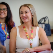 Mulher sentada em sala de aula sorri durante um encontro. Ela usa blusa clara sem mangas e crachá colorido no pescoço. Ao fundo, outra mulher de óculos também sorri. Sobre as mesas há cadernos e objetos pessoais, em ambiente de formação ou reunião.