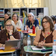Grupo de pessoas sentadas em sala de aula acompanha uma formação. Duas mulheres em primeiro plano, com crachá, óculos e cadernos, escutam atentamente. Sobre as mesas há garrafas de água, notebook e anotações. Ao fundo, estantes com livros.