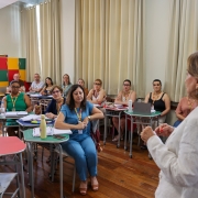 A secretária da Educação, Raquel Teixeira, fala para um grupo de pesssoas sentadas em mesas redondas. Todos têm laptops, crachás e materiais de trabalho. O local é claro, com piso de madeira, grandes janelas e paredes brancas com estante de livros.