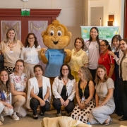 Grupo de pessoas posa para foto em ambiente interno de evento. Ao centro, um mascote sorridente com camiseta &ldquo;Mente Viva&rdquo;. Participantes est&atilde;o em p&eacute; e agachados, pr&oacute;ximos a um tel&atilde;o e banner do programa, em clima de integra&ccedil;&atilde;o.