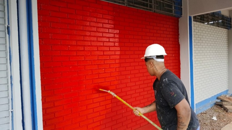 Fotografia de um homem de capacete branco e camiseta preta pintando uma parede de tijolos de vermelho vivo. Ele utiliza um rolo de pintura com extensor. &Agrave; direita, parte da parede permanece no cinza original, e as molduras da estrutura possuem detalhes em azul.