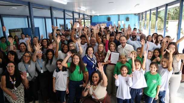 Grupo grande de estudantes, professores e autoridades reunidos em um corredor escolar iluminado, sorrindo e acenando para a c&acirc;mera. Crian&ccedil;as com uniformes escolares aparecem na frente, enquanto adultos ocupam o fundo da imagem em clima de celebra&ccedil;&atilde;o.