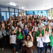 Grupo grande de estudantes, professores e autoridades reunidos em um corredor escolar iluminado, sorrindo e acenando para a c&acirc;mera. Crian&ccedil;as com uniformes escolares aparecem na frente, enquanto adultos ocupam o fundo da imagem em clima de celebra&ccedil;&atilde;o.