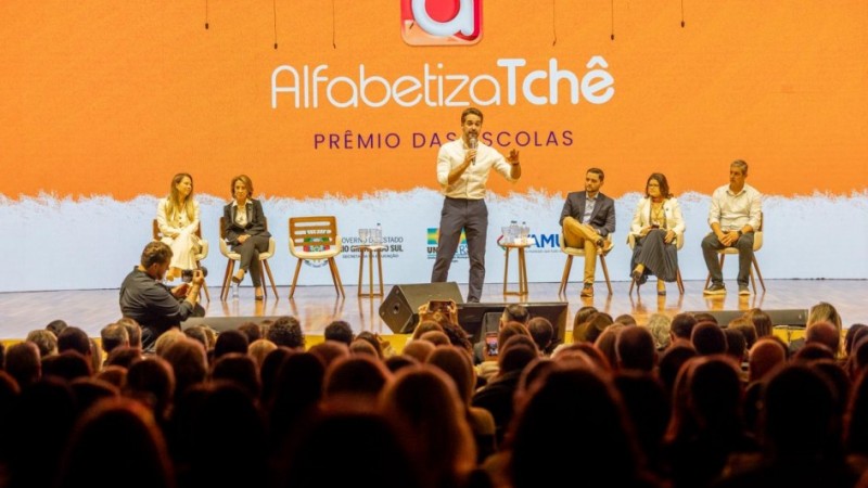 Vis&atilde;o ampla do palco durante evento, com o governador ao centro conduzindo a apresenta&ccedil;&atilde;o e outras pessoas sentadas em cadeiras alinhadas ao fundo. O painel LED exibe &ldquo;Alfabetiza Tch&ecirc; &ndash; Pr&ecirc;mio das Escolas&rdquo;, al&eacute;m de logotipos institucionais do governo estadual e parceiros. A plateia ocupa toda a &aacute;rea frontal do audit&oacute;rio.