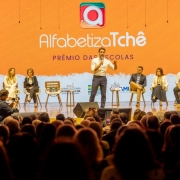 Vis&atilde;o ampla do palco durante evento, com o governador ao centro conduzindo a apresenta&ccedil;&atilde;o e outras pessoas sentadas em cadeiras alinhadas ao fundo. O painel LED exibe &ldquo;Alfabetiza Tch&ecirc; &ndash; Pr&ecirc;mio das Escolas&rdquo;, al&eacute;m de logotipos institucionais do governo estadual e parceiros. A plateia ocupa toda a &aacute;rea frontal do audit&oacute;rio.