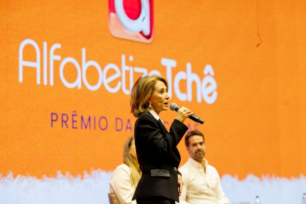 Detalhe lateral da secretária da Educação falando ao microfone no palco, posicionada diante do painel “Alfabetiza Tchê – Prêmio das Escolas”. Em segundo plano, outras pessoas permanecem sentadas em cadeiras, compondo a mesa do evento.