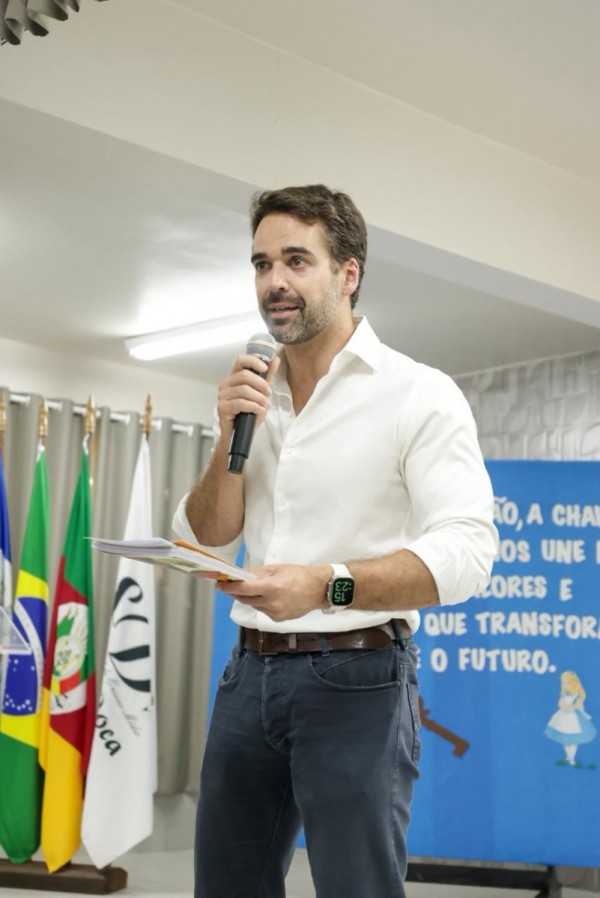 A imagem mostra o governador segurando um microfone e documentos em mãos, posicionado diante de bandeiras oficiais.