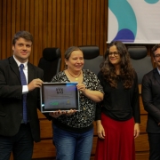 A imagem mostra um grupo de cinco pessoas alinhadas segurando um pr&ecirc;mio.