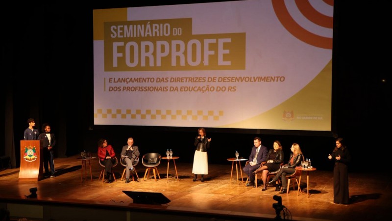 Fotografia de um palco de teatro iluminado, com um grande tel&atilde;o ao fundo onde se l&ecirc; "Semin&aacute;rio do FORPROFE e Lan&ccedil;amento das Diretrizes de Desenvolvimento dos Profissionais da Educa&ccedil;&atilde;o do RS". No palco, h&aacute; sete autoridades sentadas ou em p&eacute;, al&eacute;m de dois jovens no p&uacute;lpito e uma int&eacute;rprete de Libras.