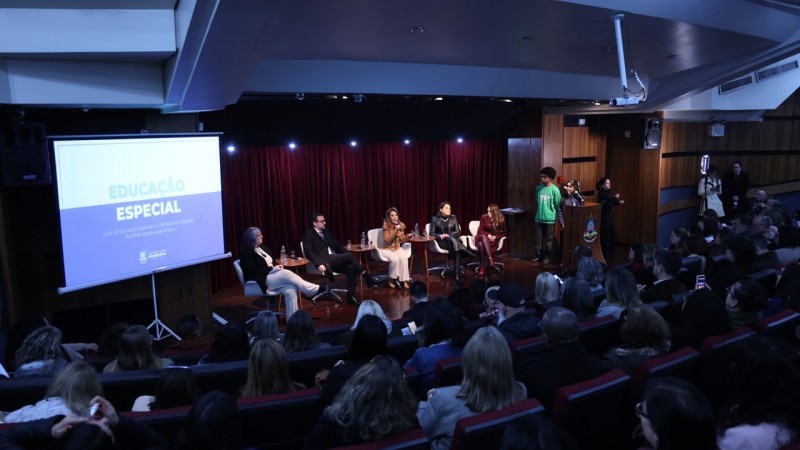 Audit&oacute;rio com plateia assistindo a uma mesa de debate sobre educa&ccedil;&atilde;o especial. No palco, cinco participantes conversam diante de uma tela que exibe o Guia da Educa&ccedil;&atilde;o Especial na Perspectiva Inclusiva da Rede Estadual de Ensino. H&aacute; int&eacute;rprete de Libras e p&uacute;blico atento.