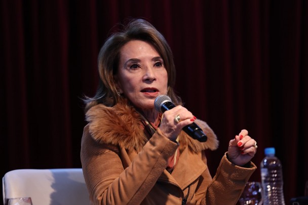 A secretária da Educação, Raquel Teixeira, está sentada em palco enquanto fala ao público. Ela veste casaco marrom com gola de pele sintética e está diante de uma cortina vermelha. Sobre a mesa ao lado, há uma garrafa de água e copos.