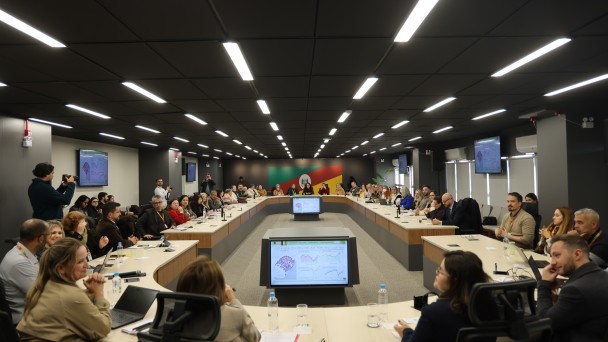Sala de reuni&otilde;es ampla com dezenas de participantes sentados ao redor de uma mesa em formato de U. Telas exibem gr&aacute;ficos e dados ao centro e nas paredes. Pessoas acompanham a apresenta&ccedil;&atilde;o, enquanto fot&oacute;grafos registram o encontro institucional.