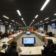 Sala de reuni&otilde;es ampla com dezenas de participantes sentados ao redor de uma mesa em formato de U. Telas exibem gr&aacute;ficos e dados ao centro e nas paredes. Pessoas acompanham a apresenta&ccedil;&atilde;o, enquanto fot&oacute;grafos registram o encontro institucional.