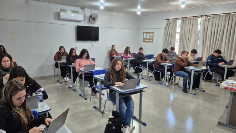Estudantes sentados individualmente em carteiras, utilizando notebooks em uma sala de aula. O ambiente tem paredes claras, janelas com cortinas, ar-condicionado, ventilador e uma televis&atilde;o fixada na parede.