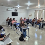 Estudantes sentados individualmente em carteiras, utilizando notebooks em uma sala de aula. O ambiente tem paredes claras, janelas com cortinas, ar-condicionado, ventilador e uma televis&atilde;o fixada na parede.