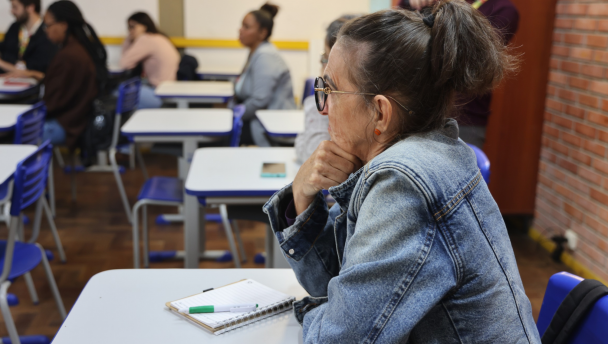 Estudante aparece sentada em sua cadeira, debruçada sobre a classe enquanto presta atenção. Ela é branca, está usando jaqueta jeans e óculos de grau com armação preta e dourada. Tem o cabelo castanho um pouco grisalho amarrado em um rabo de cavalo. 