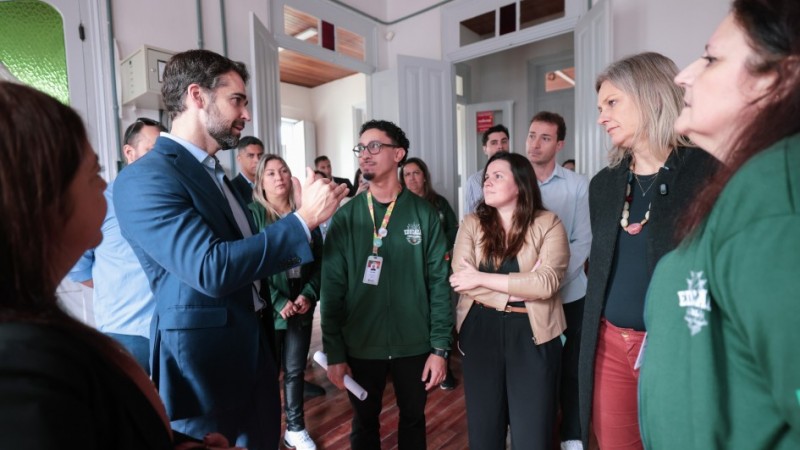 Foto de um grupo de pessoas no interior do col&eacute;gio. &Agrave; esquerda, o governador Eduardo Leite veste um terno azul e gesticula enquanto fala para os demais, que prestam aten&ccedil;&atilde;o. Entre os ouvintes, nota-se pessoas vestindo jaquetas verdes de uniforme da Rede Estadual. O local possui portas brancas e piso de madeira.