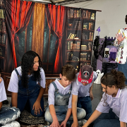 Cinco estudantes aparecem sentados em um tapete no ch&atilde;o interagindo com um livro. Uma menina de chap&eacute;u est&aacute; em p&eacute; ao lado deles. Ao fundo, um cen&aacute;rio de pano com imagem de uma estante cheia de livros e uma janela.